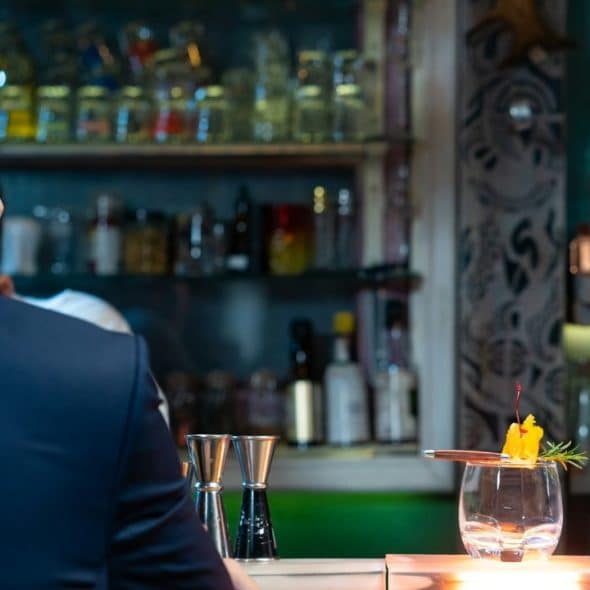 A person with a sharp haircut sits at a bar, looking at a cocktail.