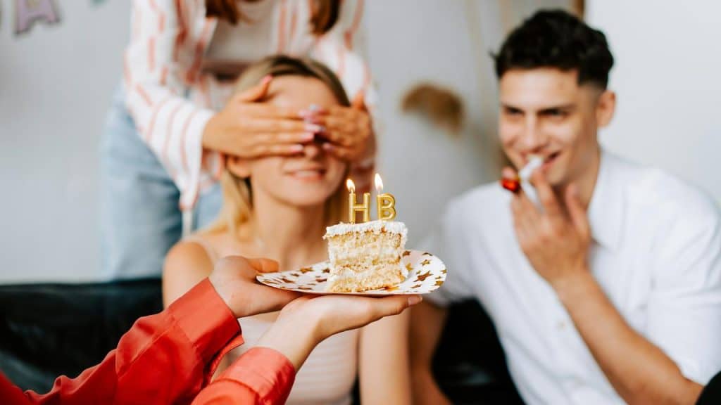 A slice of birthday cake with “HB” candles presented to a blindfolded woman.