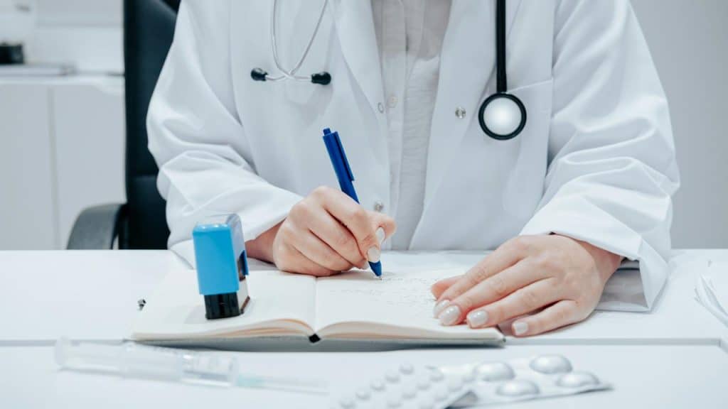 A doctor writing in a notebook with medical supplies on the desk.