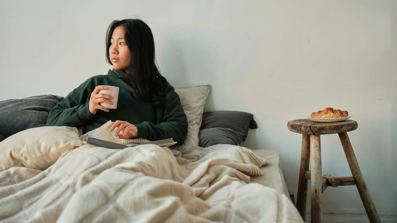 A woman sitting in bed holding a mug and an open book.