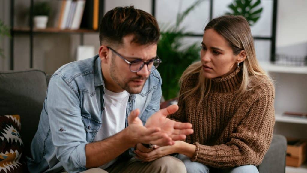 A man with glasses is talking and gesturing to a woman in a brown sweater, who looks concerned.