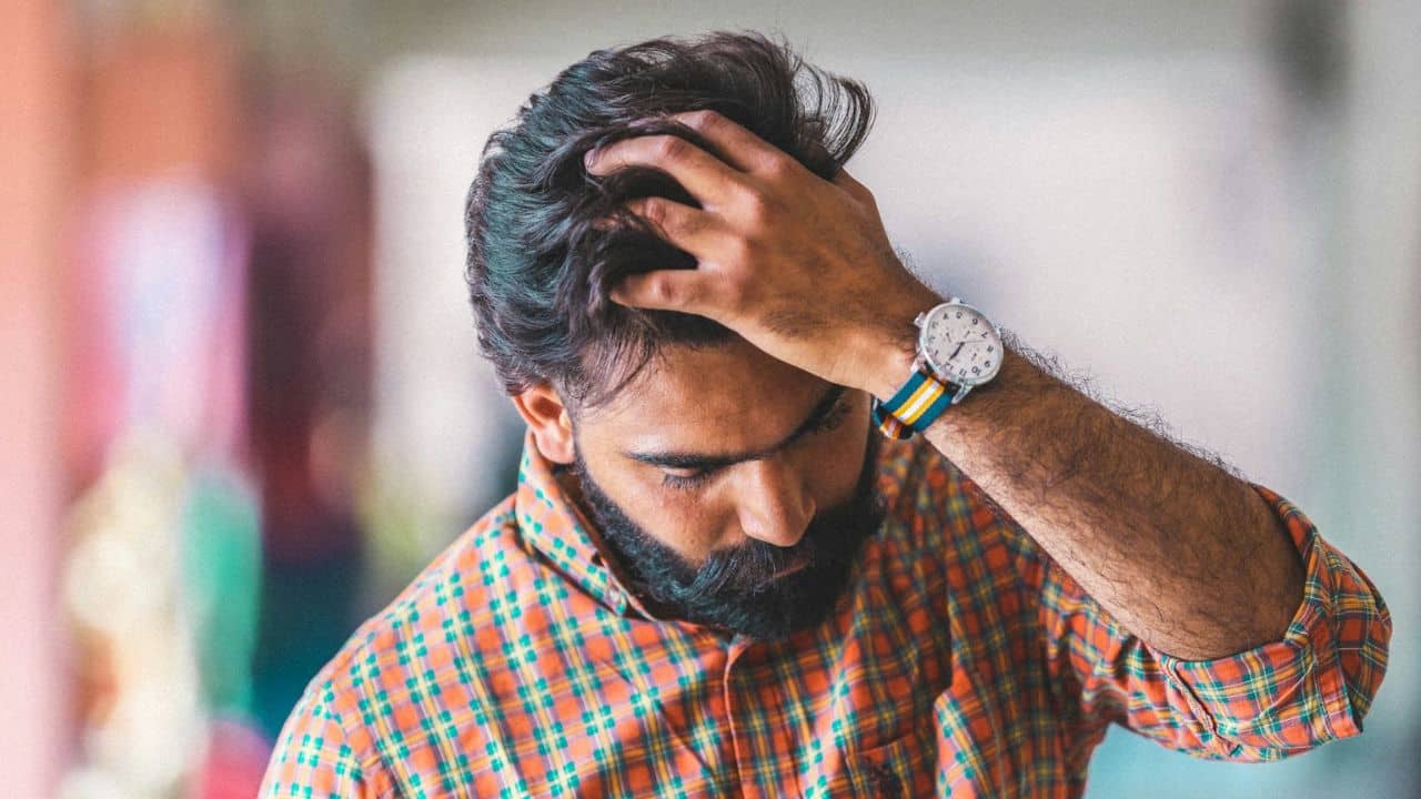 A man with a beard and a plaid shirt clutches his hair, looking down.