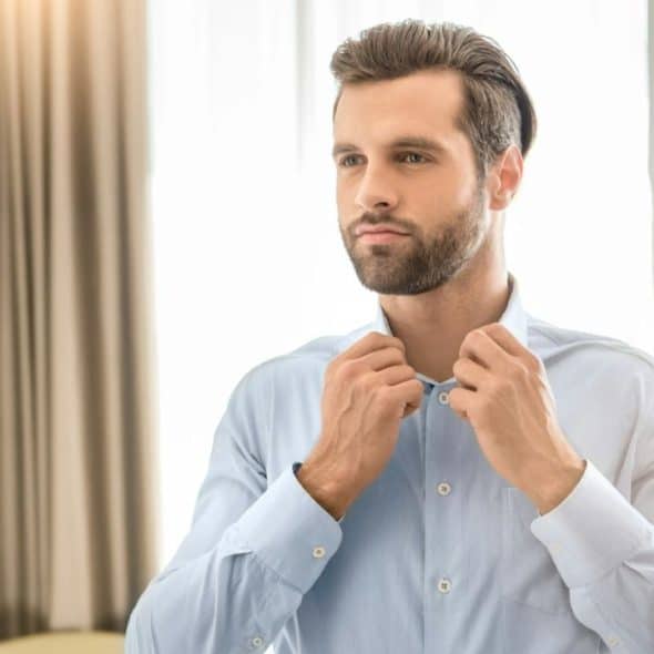 A bearded man in a light blue shirt adjusts his collar in front of a window.