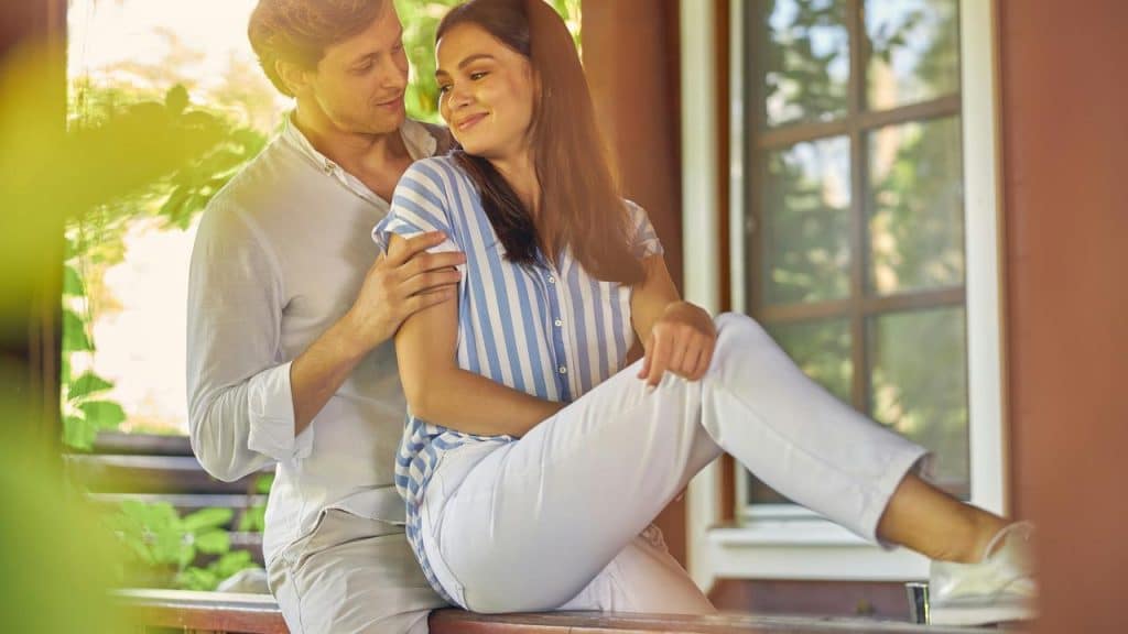 A man gently embracing a smiling woman as they sit on a porch bench.