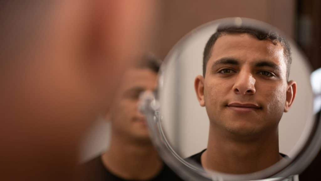 A man's face is reflected in a round mirror, with a blurred reflection of him behind it.