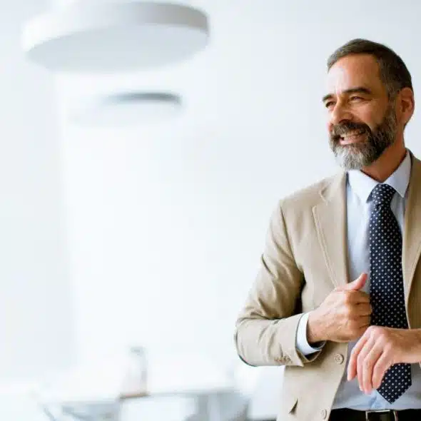 A smiling, bearded man in a beige suit and polka-dot tie looks to the right.