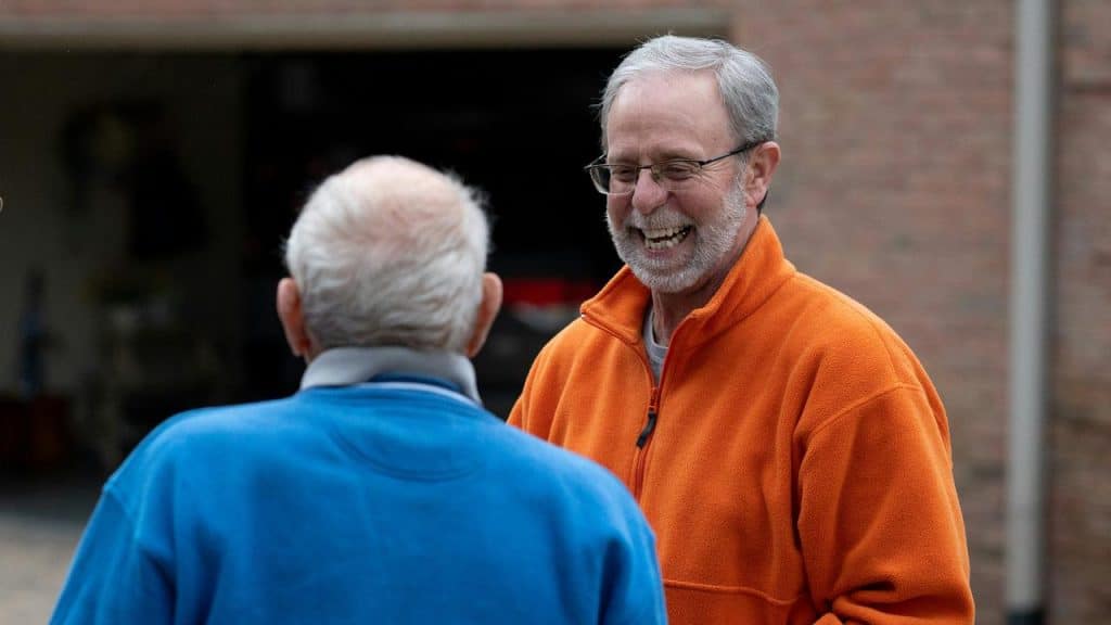 An older man in an orange fleece laughing with a friend in a blue jacket.