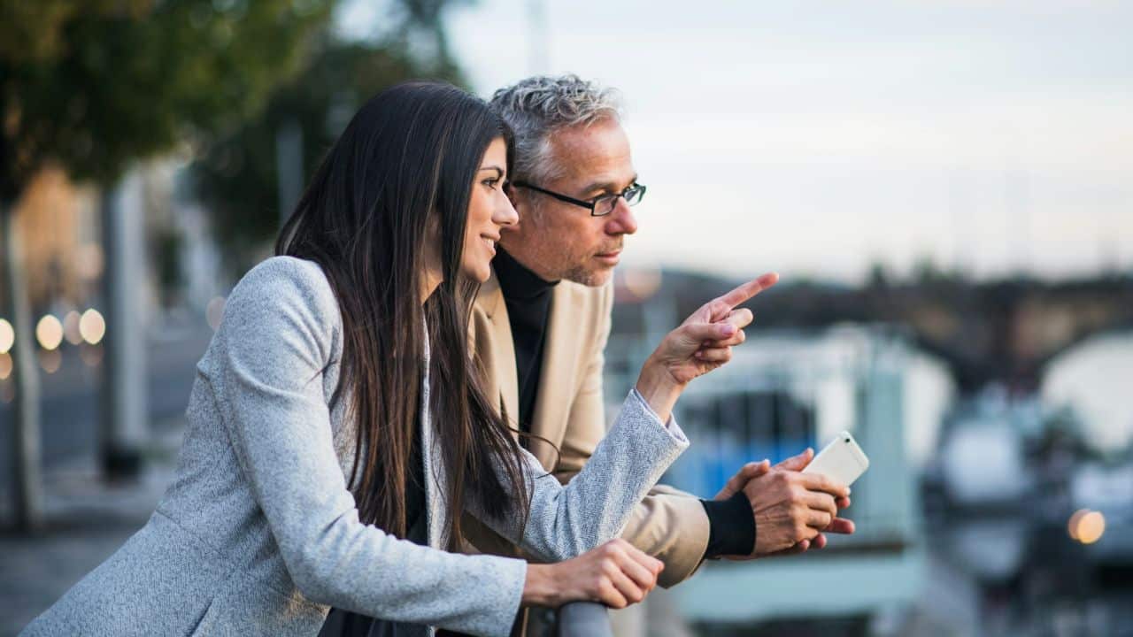 A smiling woman with long dark hair points into the distance, next to a man holding a phone.
