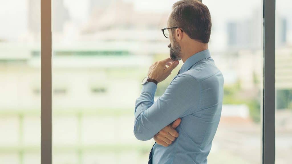 A man with glasses and a beard, wearing a light blue shirt, looks out a window.