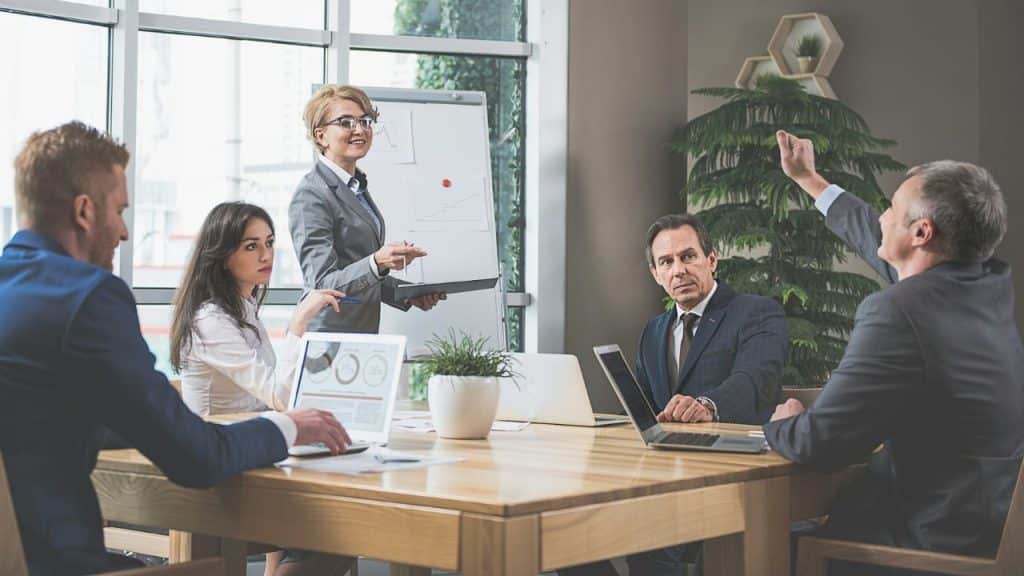 A business meeting with a woman presenting and a man raising his hand to speak.