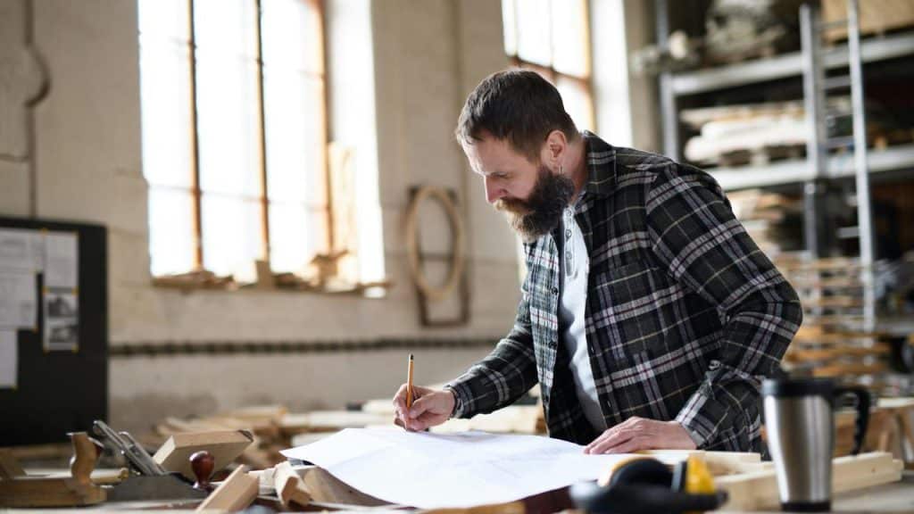 A man sketching blueprints on a workbench in a woodshop.