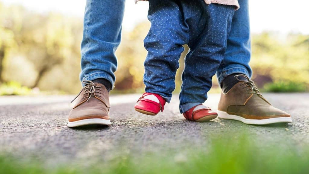 A toddler’s red shoe stepping beside an adult’s brown shoe on pavement.