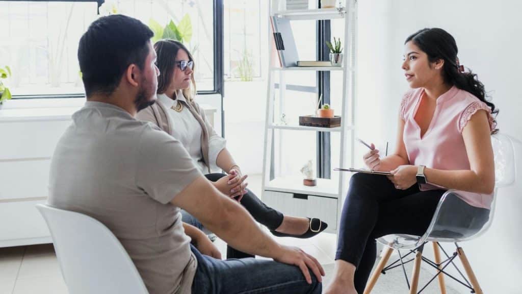A diverse couple sits facing a female therapist who is taking notes.