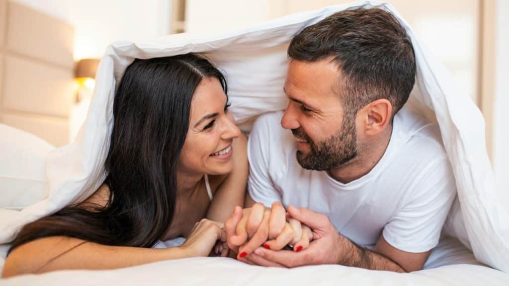 A smiling couple holding hands while lying under a white blanket in bed.