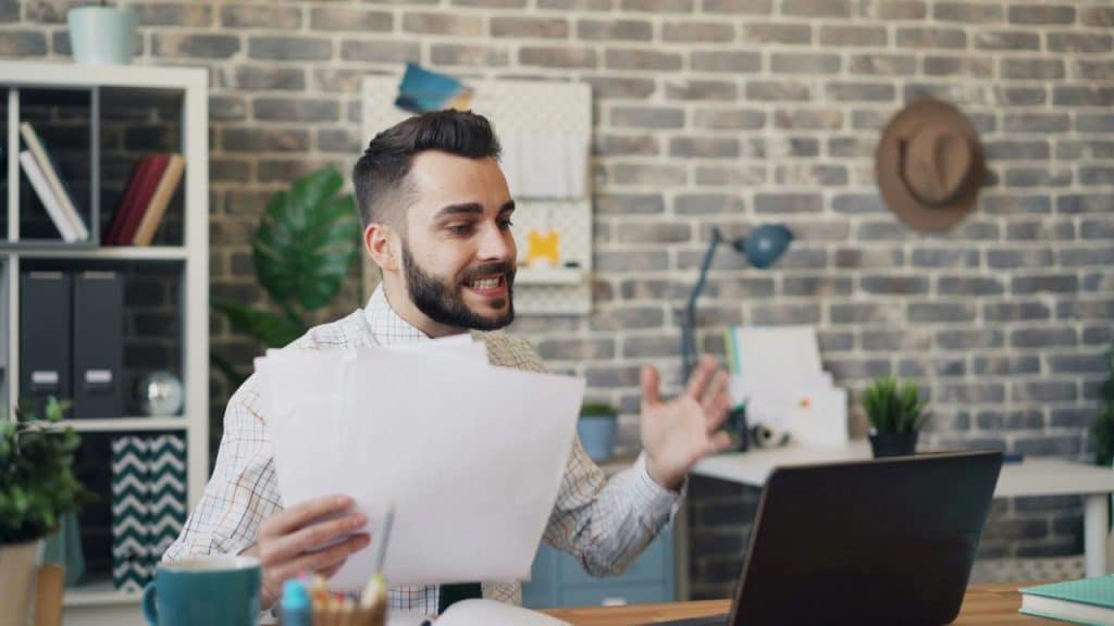 A man holding papers and angrily gesturing at his laptop.