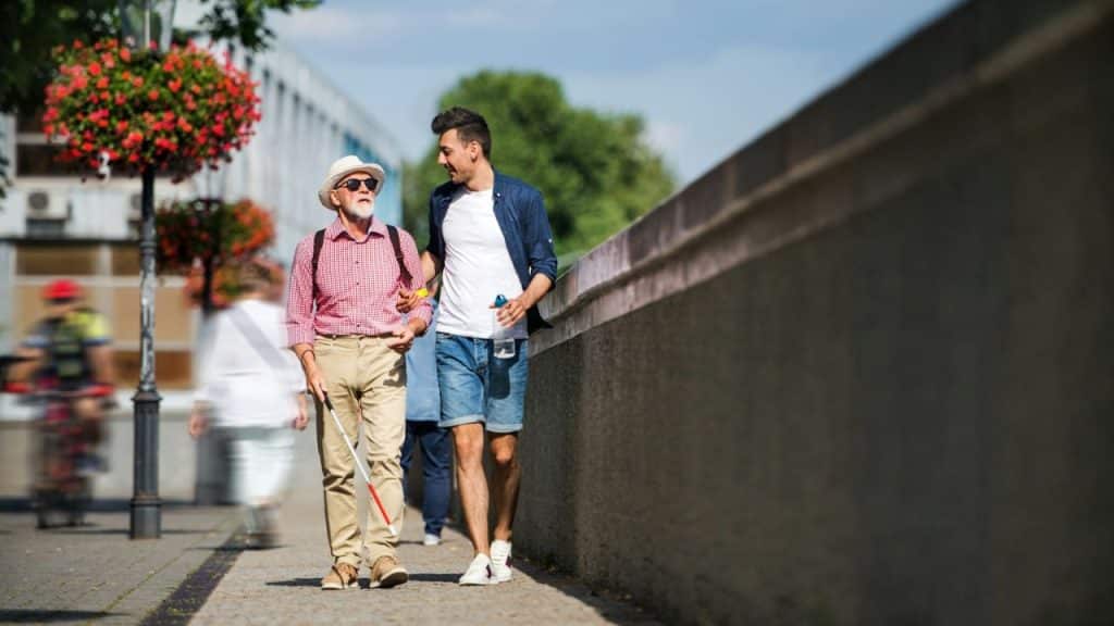 A young man helps an older man with a cane walk along a sidewalk.