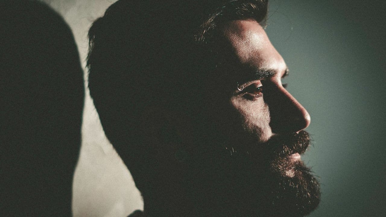 A man with a beard is depicted in profile, half-shadowed by a wall.