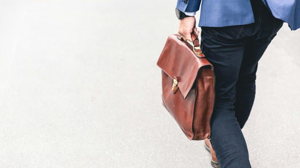 A man in a suit jacket and jeans walks carrying a brown briefcase.