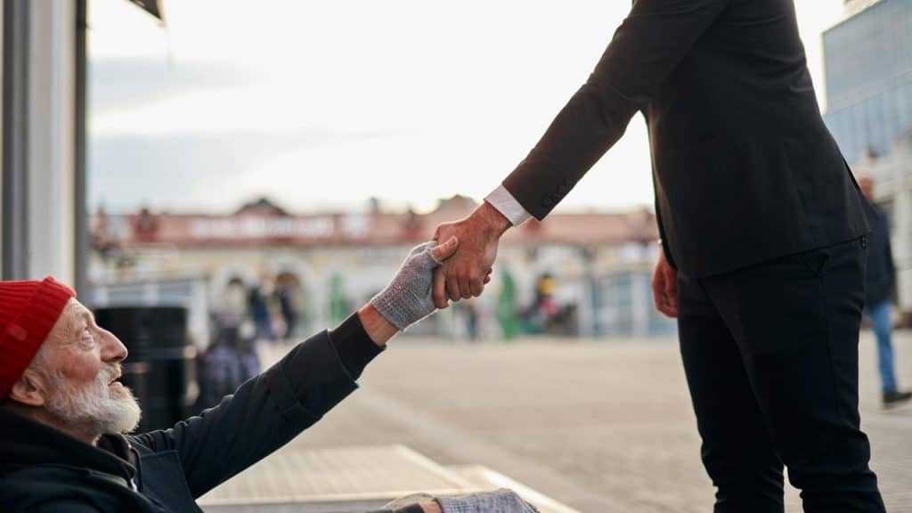 A suited man helping a homeless man up by grasping his hand.