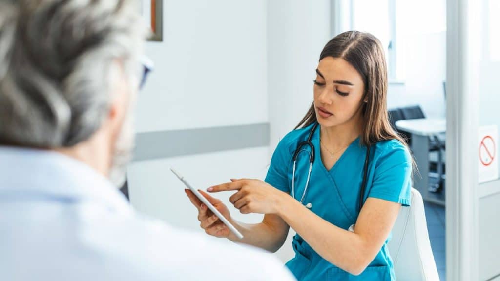 A healthcare professional in blue scrubs shows a tablet to a patient.