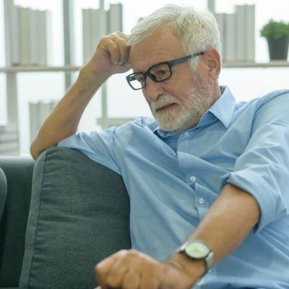A gray-haired man in a blue shirt and glasses sits on a couch, looking troubled.