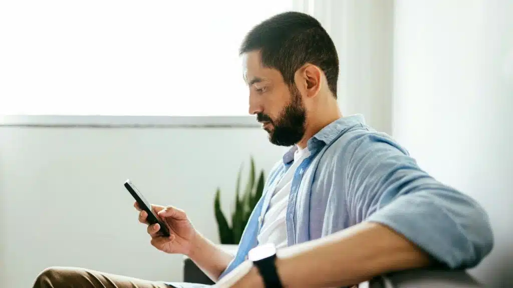 A man with a beard, wearing a light blue shirt, looks at his phone while sitting.