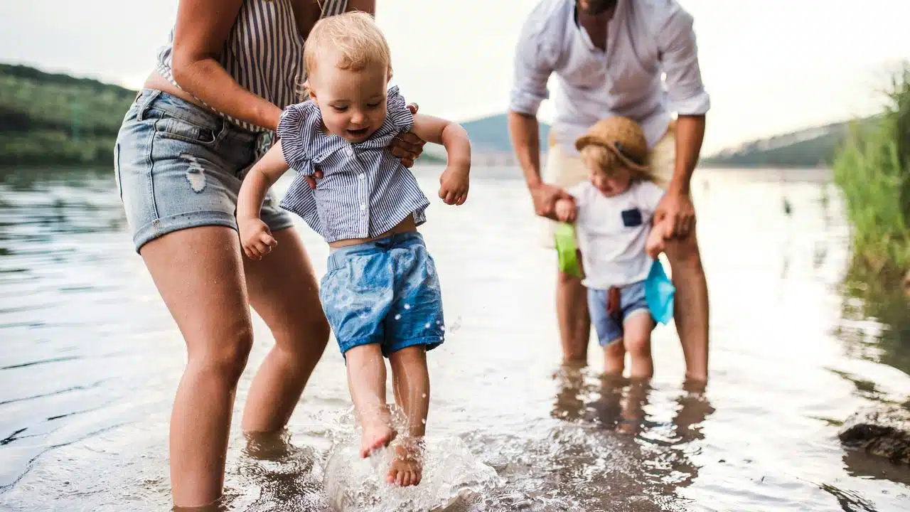 A mother and father helping two toddlers splash in shallow lake water.
