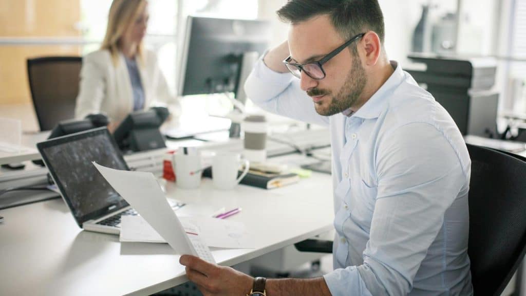 A man in glasses reading a printout at his office desk.