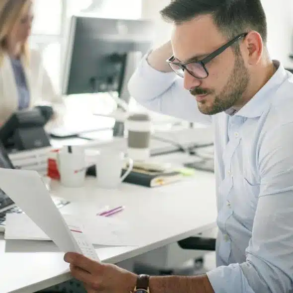 A man in glasses reading a printout at his office desk.