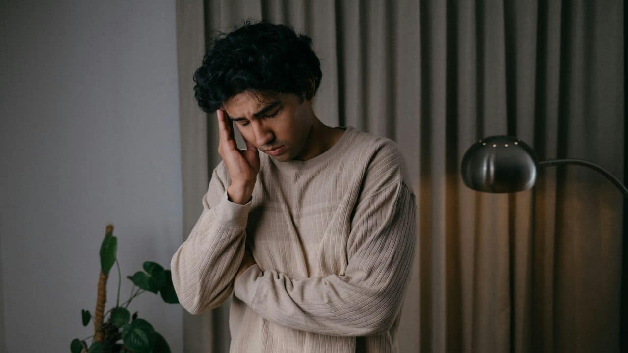 A man with curly hair stands, hand to his temple, looking downcast.