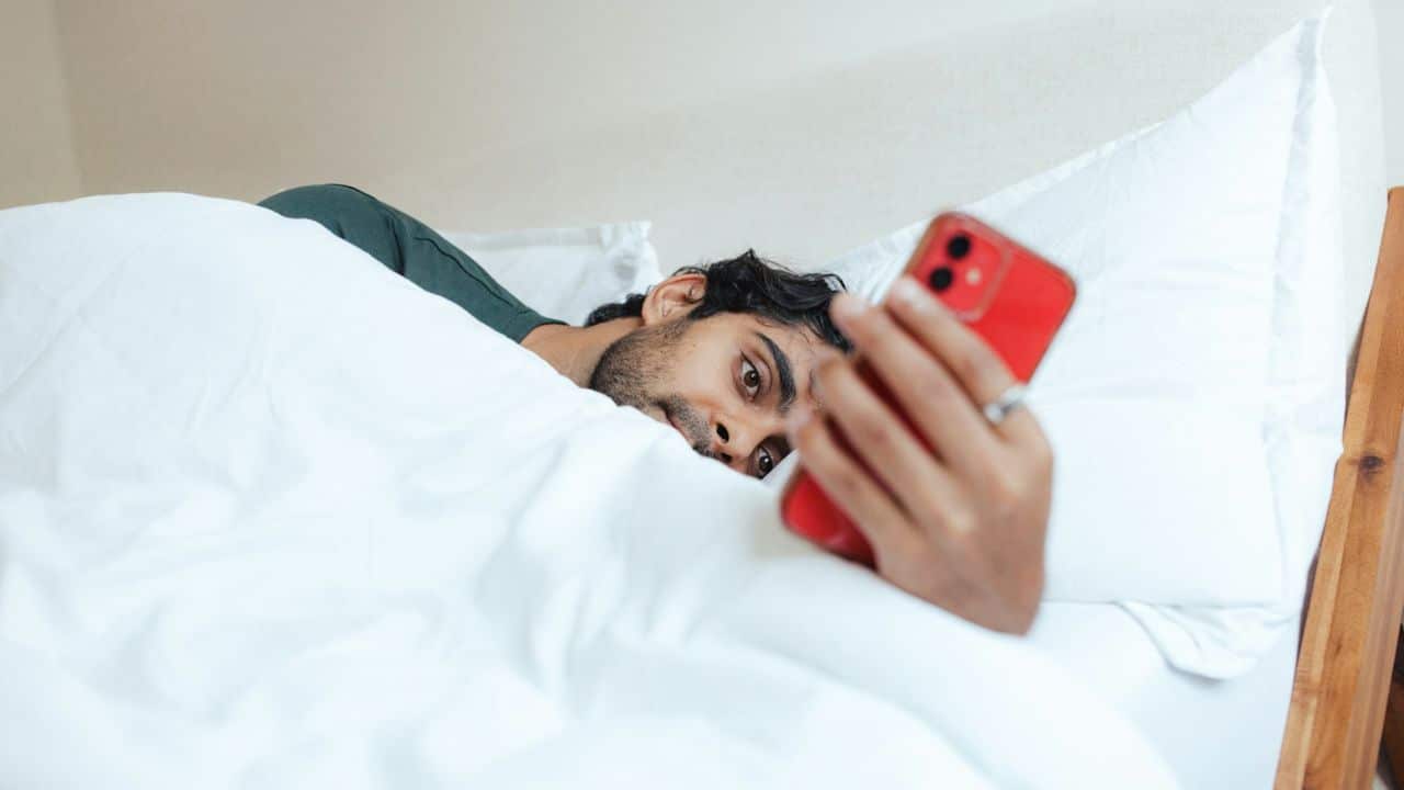 A man with a beard lies in bed under white covers, looking at a red phone.