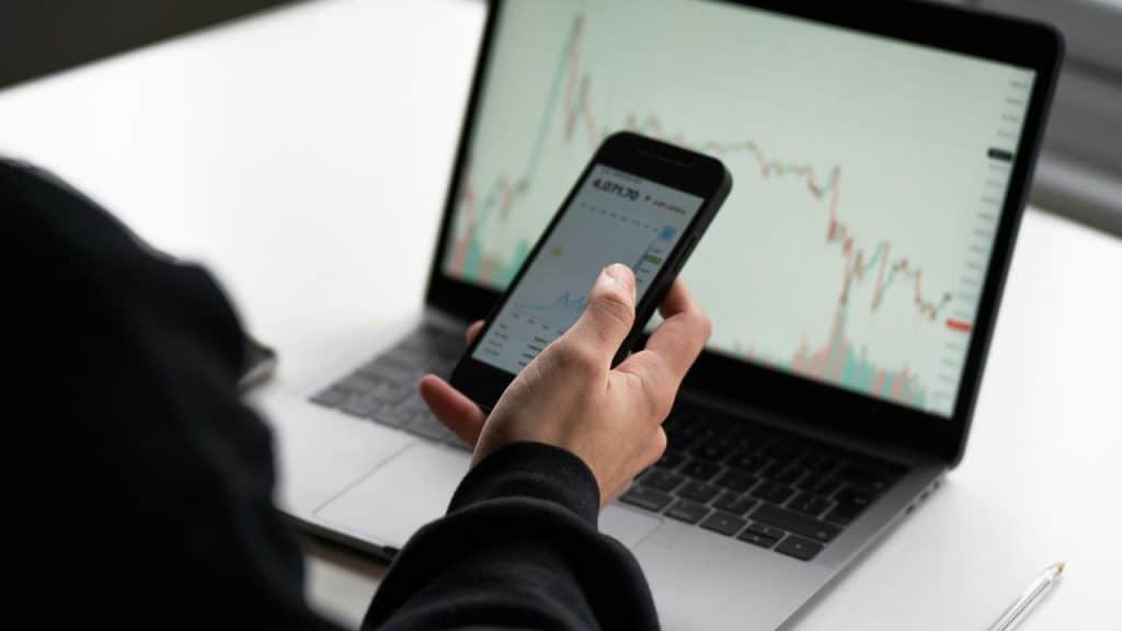 A person taking notes beside a laptop and smartphone showing stock charts.