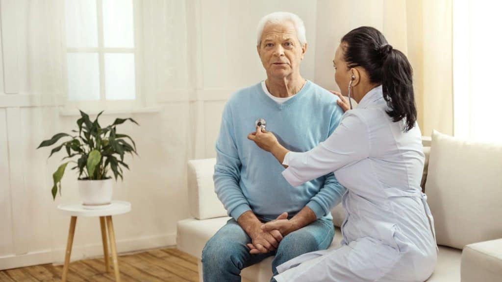 A doctor in a white coat uses a stethoscope to examine a senior man in a blue sweater.