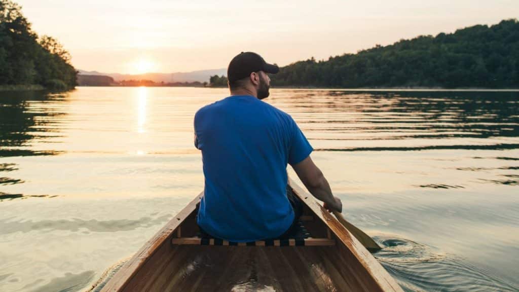 A man in a blue shirt and cap paddles a canoe on a lake at sunset.