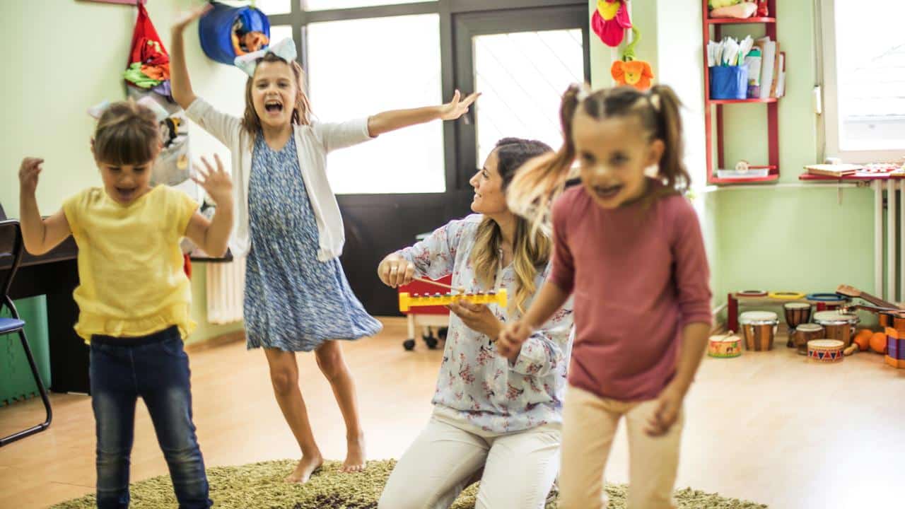 A teacher playing a xylophone as children dance around in a classroom.