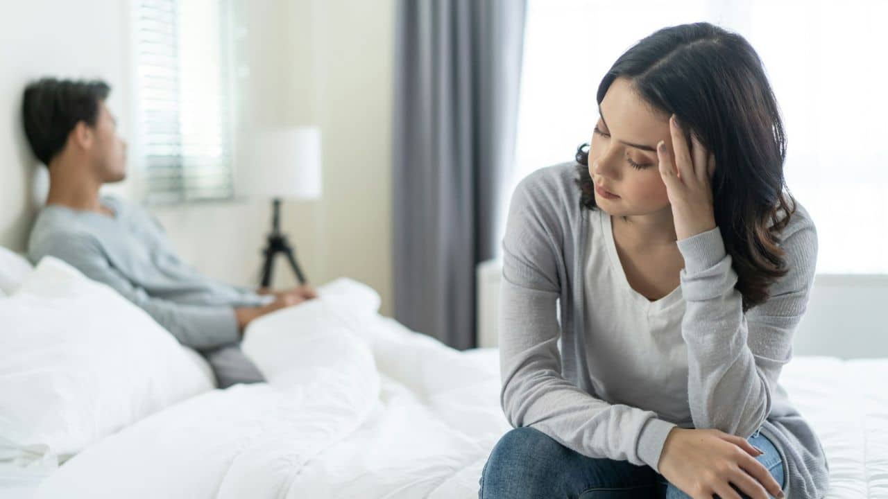 A woman sits on a bed with her hand on her head, looking away from a man in the background.