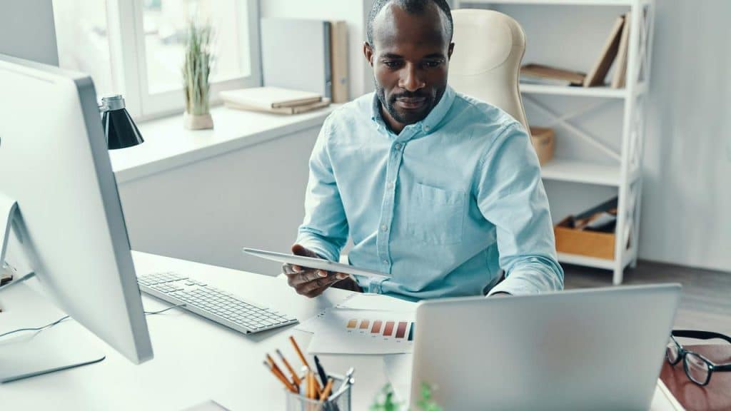 A man seated at a desk, holding a tablet and looking at a laptop.