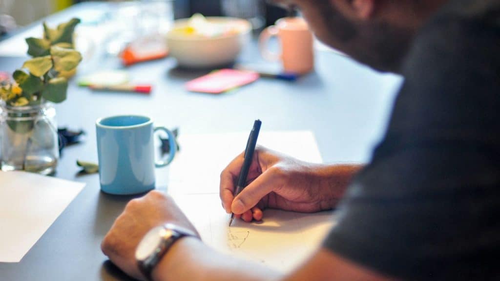 A person with a watch on their wrist writes on paper, with a blue mug nearby.