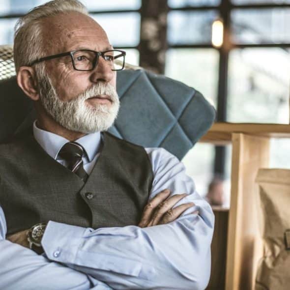 A grey-bearded man in a shirt, tie, and vest sits with arms crossed, looking thoughtful.
