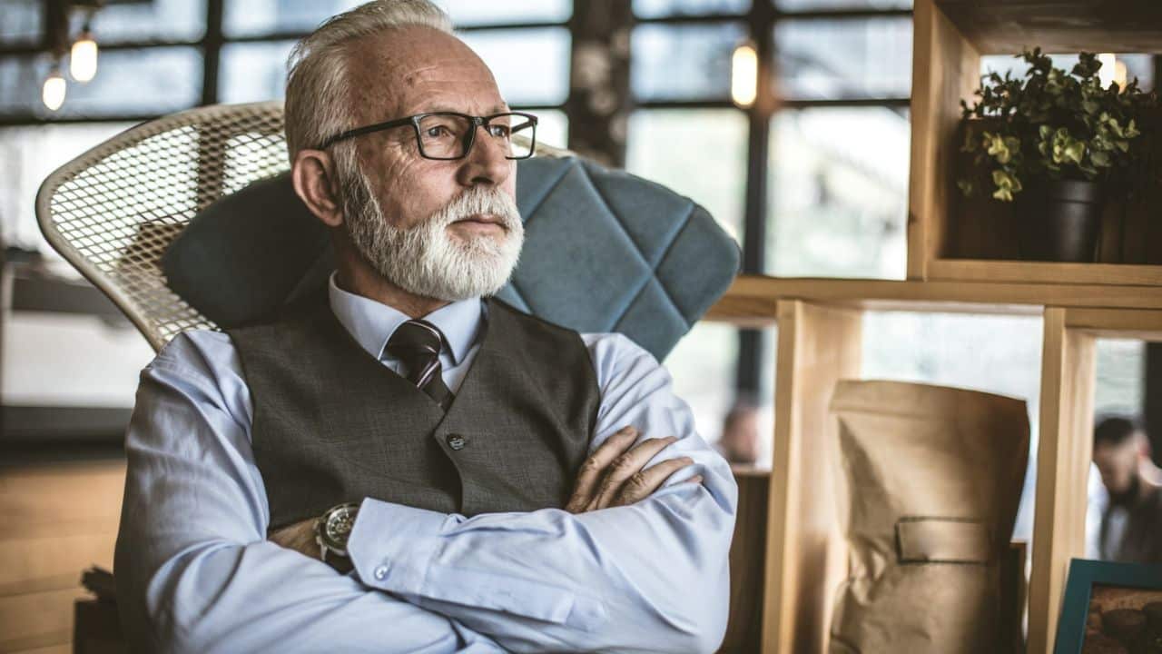 A grey-bearded man in a shirt, tie, and vest sits with arms crossed, looking thoughtful.
