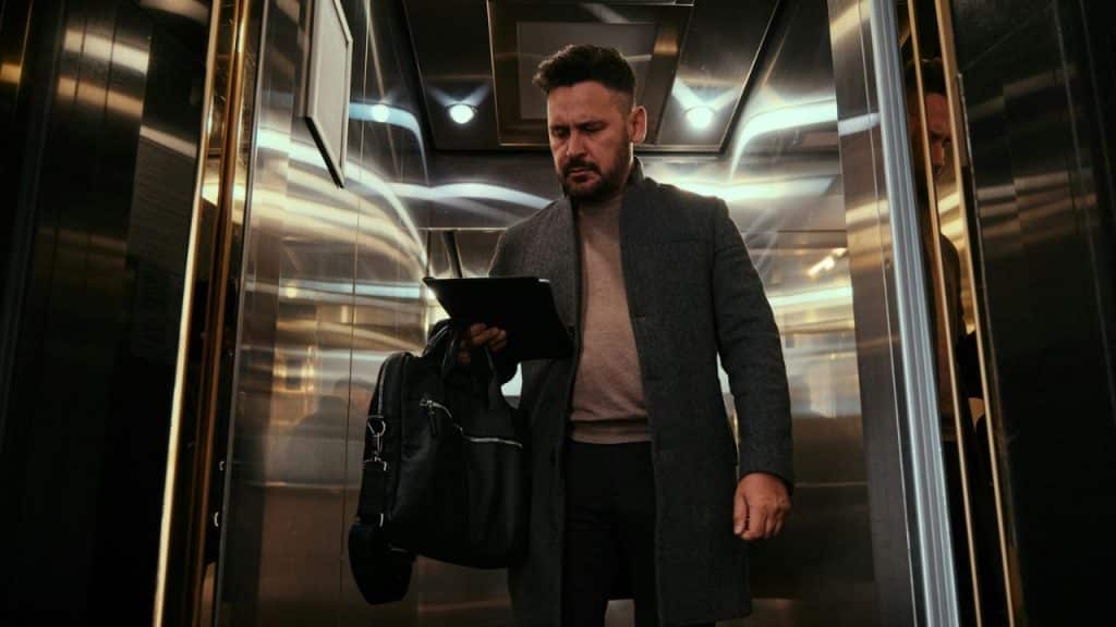 A man with a beard and dark hair, wearing a gray coat and holding a tablet and bag, stands in an elevator.