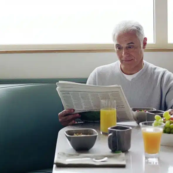A smiling, grey-haired man reads a newspaper at a diner booth, with breakfast and fruit on the table.