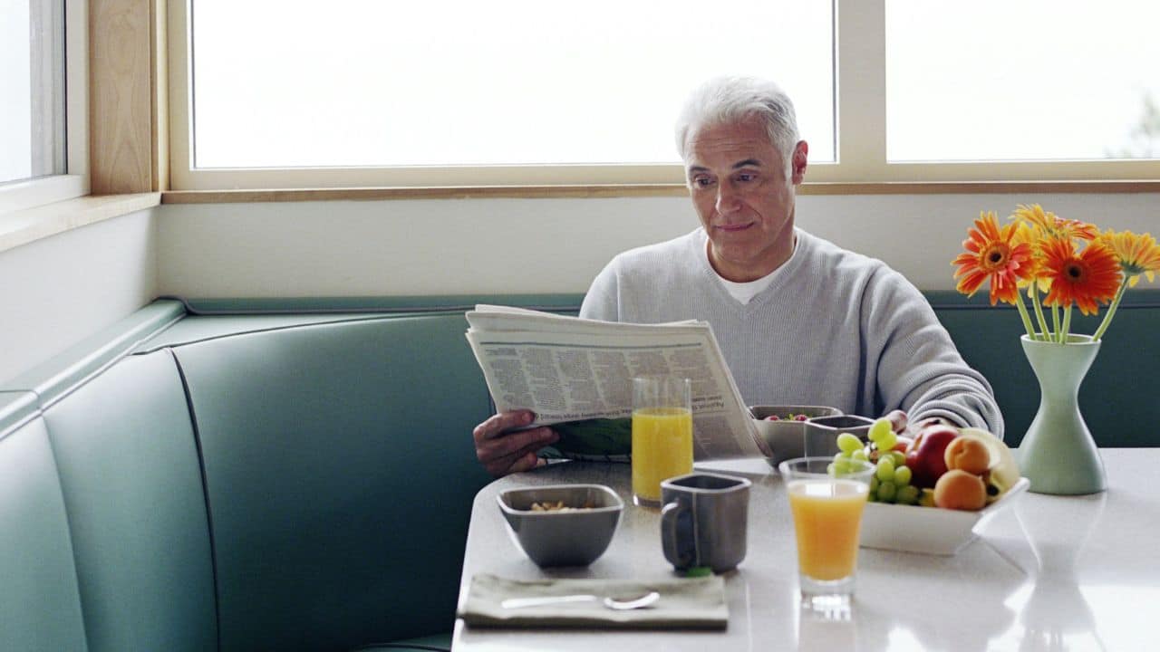 A smiling, grey-haired man reads a newspaper at a diner booth, with breakfast and fruit on the table.