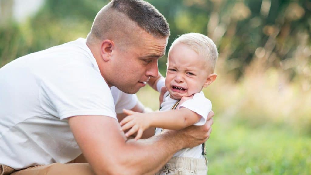 A man holding a crying toddler in a grassy field.