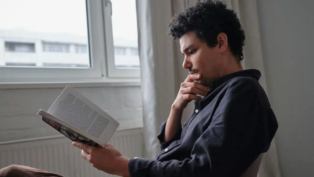 A man with dark curly hair reads a book, resting his chin on his hand.