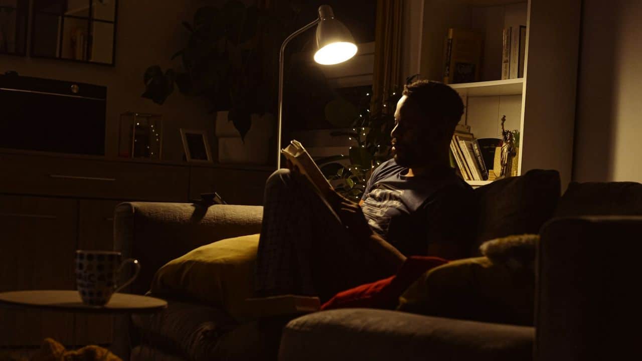 A man sits on a sofa, reading a book under the light of a floor lamp.