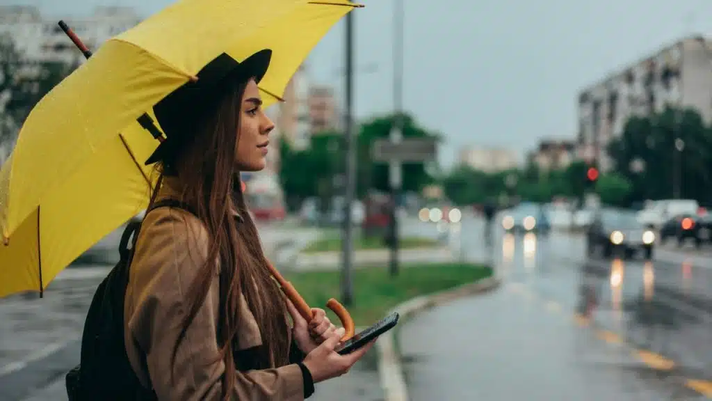 A young woman holds a yellow umbrella and a phone on a rainy street.