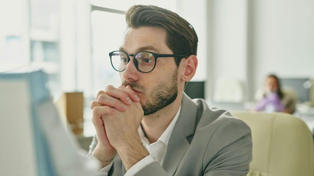 A man with glasses and a beard, in a suit, clasps his hands and looks intently to the side.