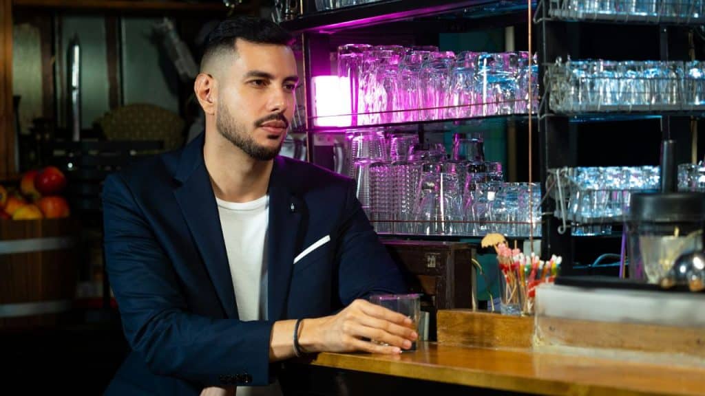 A man with a beard in a navy suit sits at a bar, holding a drink.