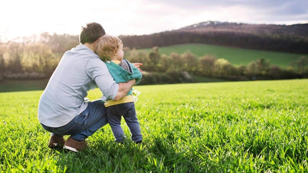 A father crouches in a green field holding his toddler.